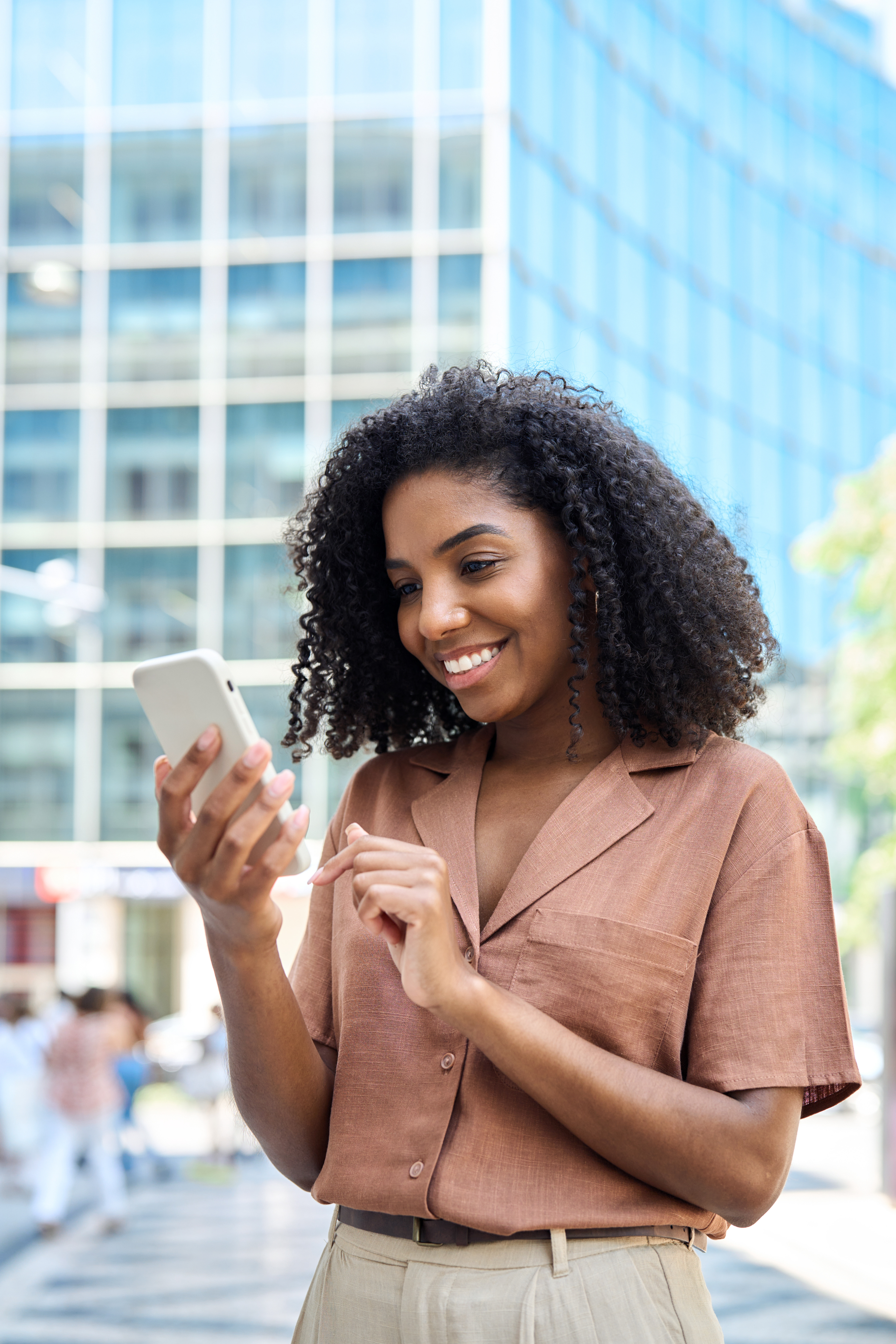 Young woman smiling outdoors while checking her phone, feeling confident about her finances.